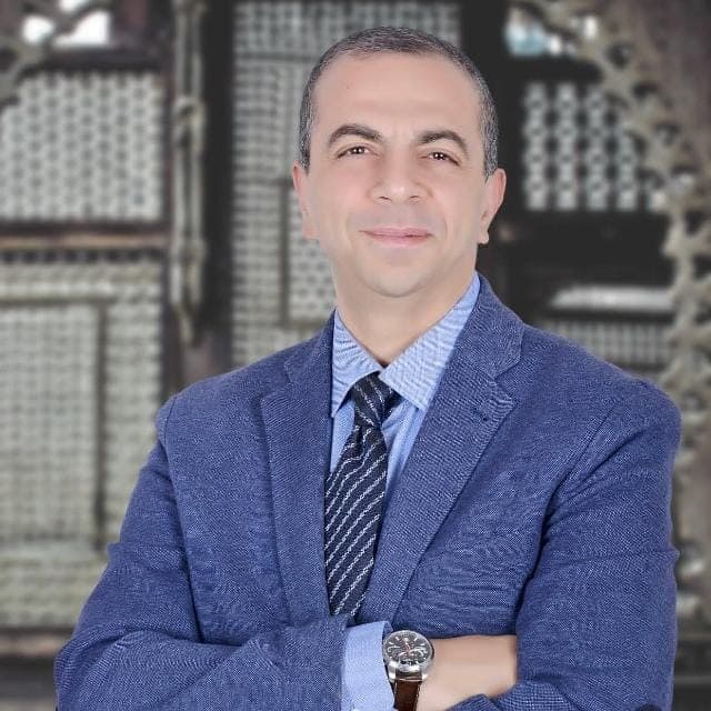 Smiling man in a blue textured blazer and tie with arms crossed against blurred background.